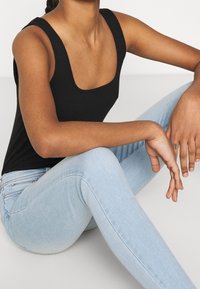 Svart ribbad linne med breda axelremmar som kombineras med ljusblå slim-fit jeans. Modellen sitter i en avslappnad pose som visar hudtonskillnader.