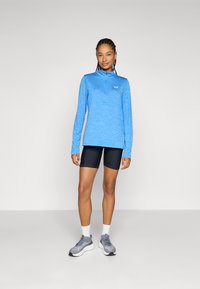 Hellblaues Langarmoberteil mit halbem Reißverschluss, kombiniert mit schwarzen Radlerhosen. Das Model trägt graue Sportschuhe mit weißen Socken.