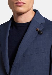 Nahaufnahme eines Mannes, der ein navyblaues strukturiertes Sakko über einem schwarzen Strickpullover trägt, mit einem kleinen Knopfetikett am Revers.