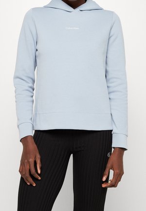 Osoba ubrana w jasnoniebieską bluzę z kapturem Calvin Klein oraz czarne, prążkowane legginsy z logo CK na udzie, stojąca na neutralnym tle.