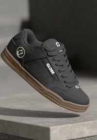 Black skate shoes with textured synthetic material, featuring a brown rubber sole, embossed logo, and white branding accents.
