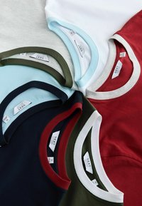 Pino taitettuja crew neck -t-paitoja eri väreissä, mukaan lukien punainen, valkoinen, laivastonsininen, vaaleansininen ja oliivinvihreä, joissa brändilaput näkyvissä kauluksissa.