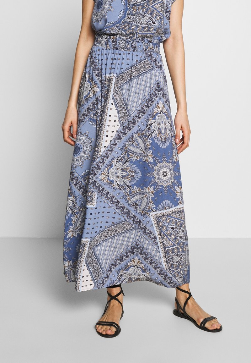 Femme portant une jupe longue bleue à motifs floraux et géométriques, associée à des sandales plates noires à brides.