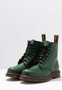 Green leather ankle boots with a smooth finish, black laces, and yellow loop tags. Features a contrasting orange stitching detail and a durable sole.