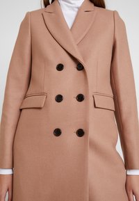 Dobbeltbrystet frakke i en blød beige stof. Har notched kraver, to sidelommer og sorte knapper til lukning. Glat tekstur.