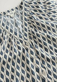 Camicia button-up realizzata in tessuto leggero, con uno sfondo crema decorato da un motivo di foglie blu navy e piccoli bottoni rotondi.