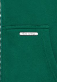 Grøn zip-up hoodie med en frontlomme og et metallisk mærke med teksten "bazix republic". Glat tekstur med subtile syninger.