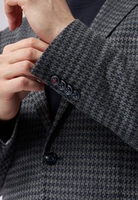 Grau und marinefarbener Hahnentritt-Blazer aus strukturiertem Stoff, mit vier schwarzen Knöpfen mit rotem Akzent und einem Reverskragen.