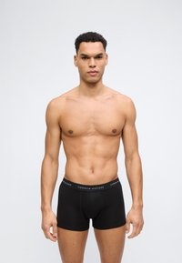 Άνδρας χωρίς μπλούζα με κοντά κατσαρά μαλλιά, φοράει μαύρα εσώρουχα Tommy Hilfiger, μπροστά σε απλό φωτεινό φόντο.