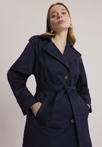 Granatowy trench w lekkim materiale, z szerokim kołnierzem, dwiema przednimi kieszeniami, związanym paskiem w talii i zapięciem na guziki.