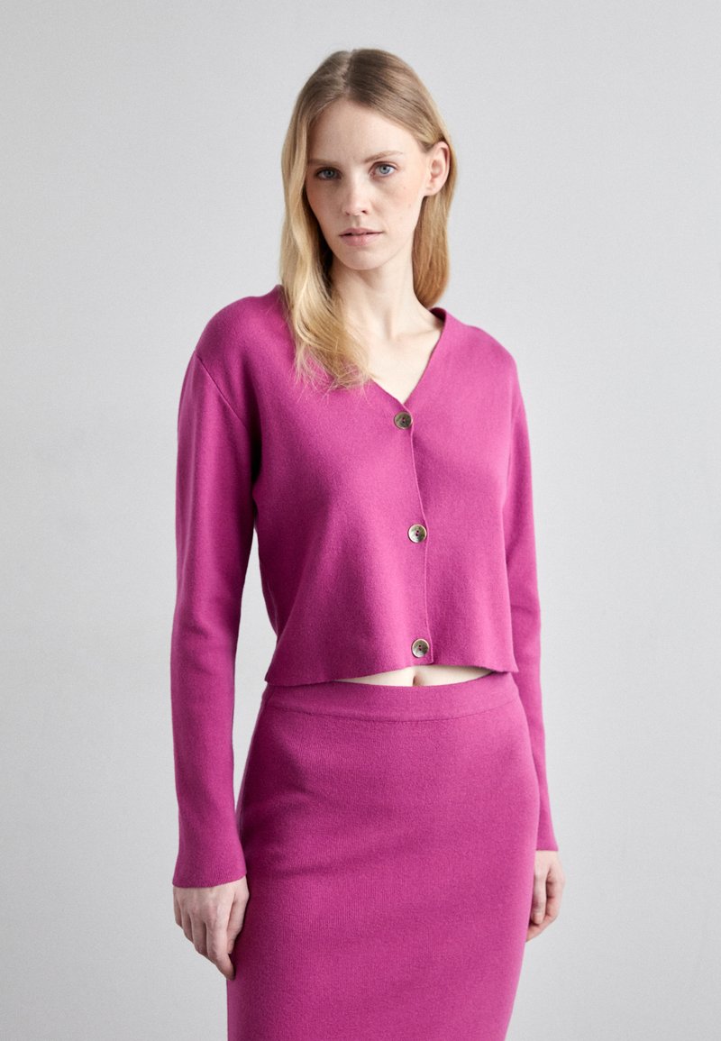 Fuchsia verkürzter Cardigan aus weichem Material mit V-Ausschnitt, einem einzelnen Knopfverschluss und langen Ärmeln, kombiniert mit einem passenden, figurbetonten Rock.
