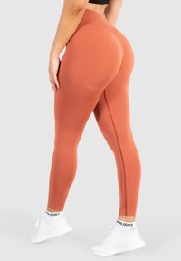 Vysoko vytažené legíny v barvě rezavě oranžové vyrobené z elastického materiálu, charakterizované hladkým texture a bočními švy. Vybavené bílými sportovními botami.