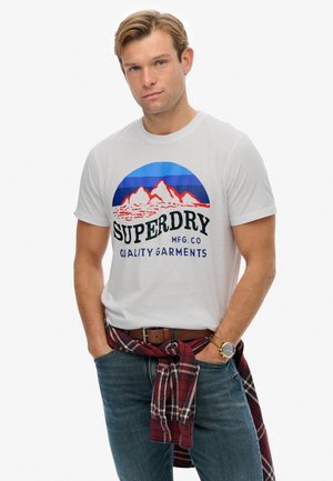 White cotton t-shirt with blue and red mountain graphic, "SUPERDRY" logo in bold black text, paired with denim jeans and a red plaid shirt tied at the waist.