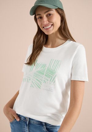 Femme souriante portant une casquette verte, un t-shirt blanc avec des motifs de feuilles vert clair et un texte, ainsi qu'un jean bleu, debout avec une main dans la poche.