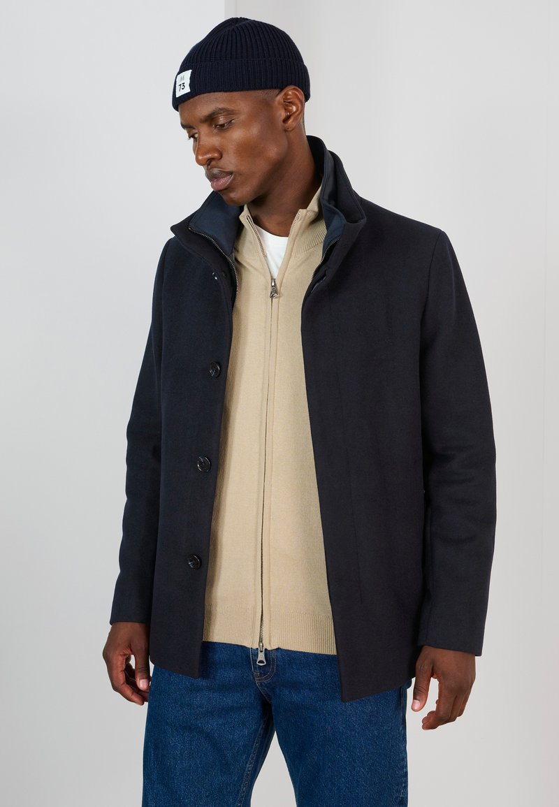 Mörk marinblå kappa med en strukturerad krage, med knappstängning. Buren över en beige zip-up tröja och blå jeans, med en stickad mössa.