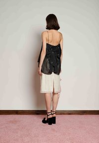 Γυναίκα που στέκεται δίπλα στον τοίχο, φορώντας μαύρο αμάνικο τοπ με μεγάλο φιόγκο στην πλάτη, κρεμ φούστα μέχρι τα γόνατα με σκίσιμο και μαύρα σανδάλια με τακούνι.