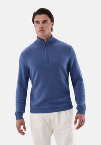 Blauer Pullover mit Reißverschluss und rundem Halsausschnitt aus weichem, strukturiertem Stoff. Mit gerippten Bündchen und Saum sowie einem modernen Schnitt ausgestattet. Kombiniert mit hellen Hosen.