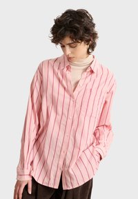 Pink collared shirt featuring vertical white stripes, button closure, and a chest pocket. The fabric has a textured appearance.
