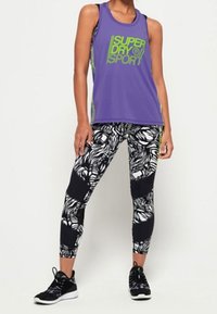 Top sportivo viola con logo verde, abbinato a leggings neri e bianchi con motivo floreale. Include scarpe da ginnastica nere.