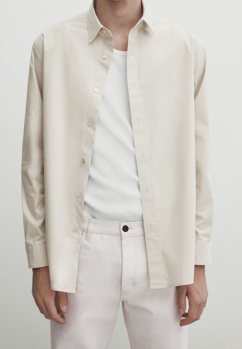 Jasno beżowa koszula zapinana na guziki z bawełny, noszona rozpięta na białym topie na ramiączkach, zestawiona z jasnoróżowymi spodniami. Prosty design, klasyczny kołnierzyk.