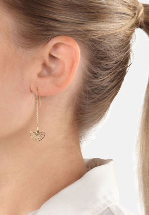 Donna con capelli biondi raccolti, indossa un delicato orecchino d'oro a forma di ventaglio appeso a una sottile catena.