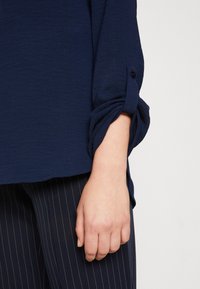 Main reposant sur le côté, portant une chemise bleu marine à manches longues avec un revers roulé et un pantalon foncé à fines rayures.