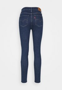 Jean skinny bleu foncé avec deux poches arrière, coutures contrastées, un patch en cuir de marque sur la ceinture et une étiquette de marque rouge sur la poche droite.