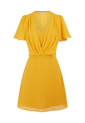 Femme portant une robe courte jaune avec un devant plissé et des manches flottantes, debout devant un fond blanc uni.
