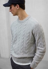 Suéter gris de punto trenzado con puños y dobladillo acanalados, con un escote redondo. Lleva encima una camisa blanca, exhibido en un modelo masculino.