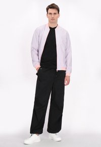 Lichtpaarse bomberjack met roze accenten, over een zwart T-shirt en losse zwarte broek; gedragen met witte sneakers.