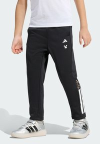 Dítě oblečené v černých kalhotách Adidas s bílými pruhy a pixelovým designem, doplněné stříbrno-černými teniskami Adidas a šedými ponožkami.