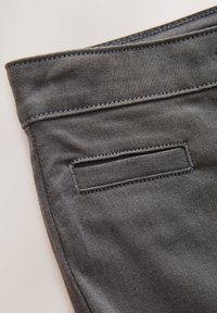 Tmavohnedý denimový materiál s hladkými švami, malou zadnou vreckou a jemnou textúrou. Pásik je mierne zatočený.