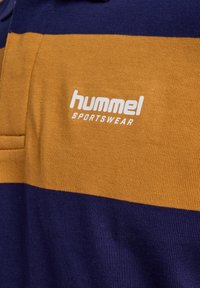Close-up van mosterdgele en marineblauwe gestreepte sportstof met wit "hummel SPORTSWEAR" logo op de borst.