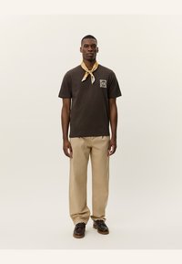 Camiseta de algodón marrón con un diseño de bolsillo en el frente, combinada con pantalones beiges y una bufanda amarilla con estampado atada al cuello. Calzado marrón.