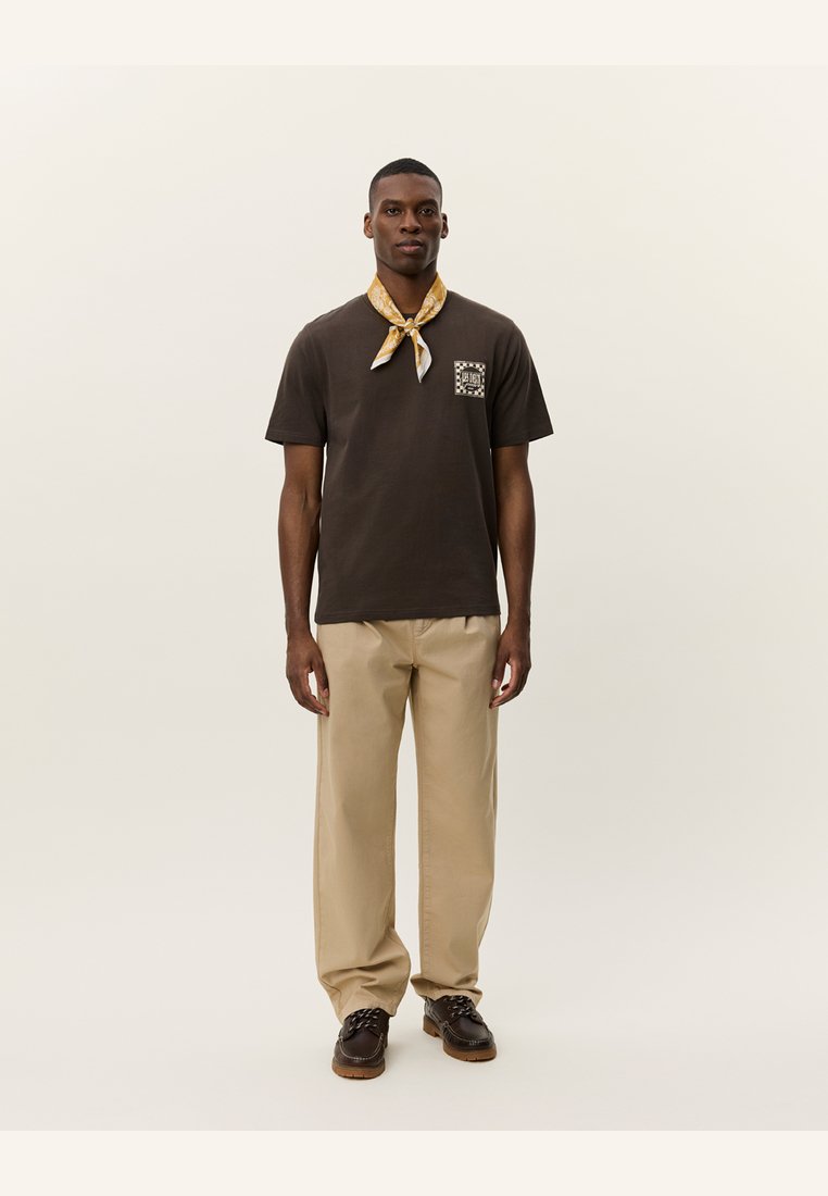 Camiseta de algodón marrón con un diseño de bolsillo en el frente, combinada con pantalones beiges y una bufanda amarilla con estampado atada al cuello. Calzado marrón.