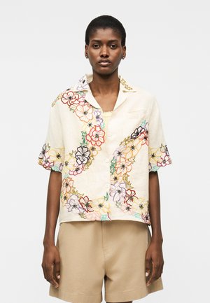 Γυναίκα που φοράει κοντομάνικο, κρεμ πουκάμισο με πολύχρωμη κεντητή λουλουδιού και μπεζ σορτς, πάνω σε απλό φόντο.