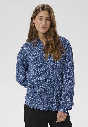 Chemise bleue à boutons avec un motif floral, présentant une coupe décontractée, des manches longues et une texture lisse. Un pantalon noir est également visible.