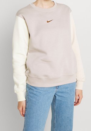 Personne portant un sweat-shirt Nike beige et crème avec un logo orange et un jean bleu, debout devant un fond uni.