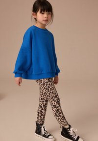 Heller blauer Oversized-Pullover, gemusterte Leopardenleggings und schwarze Sneakers mit hohem Schaft. Glattes Material, lässige Passform und kontrastierende Designs.