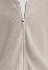 Tissu tricoté beige avec une fermeture éclair argentée partiellement fermée, révélant un tissu blanc en dessous.