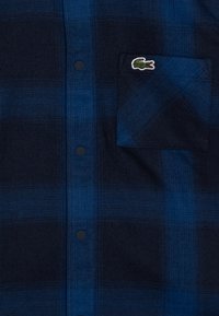 Camicia a quadri blu navy con accenti blu scuri, colletto classico, bottoni neri e taschino a sinistra con un logo ricamato verde.