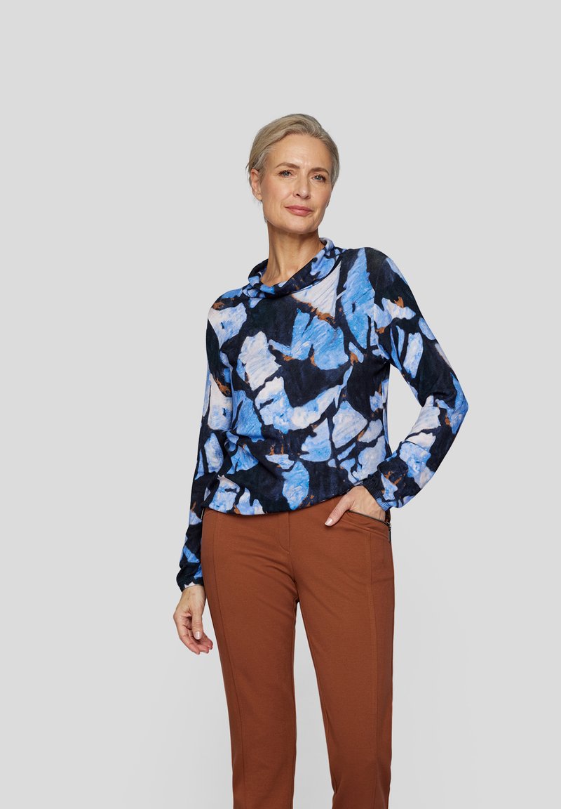 Langärmliges Oberteil mit blau-weißem Muster und einem Schalkragen, kombiniert mit braunen Hosen; glatte Textur, lockere Passform und sichtbare Taschenverarbeitung.