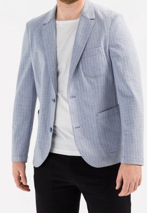 Mężczyzna w jasnoniebieskiej marynarce w prążki, białym T-shircie i czarnych spodniach na białym tle.
