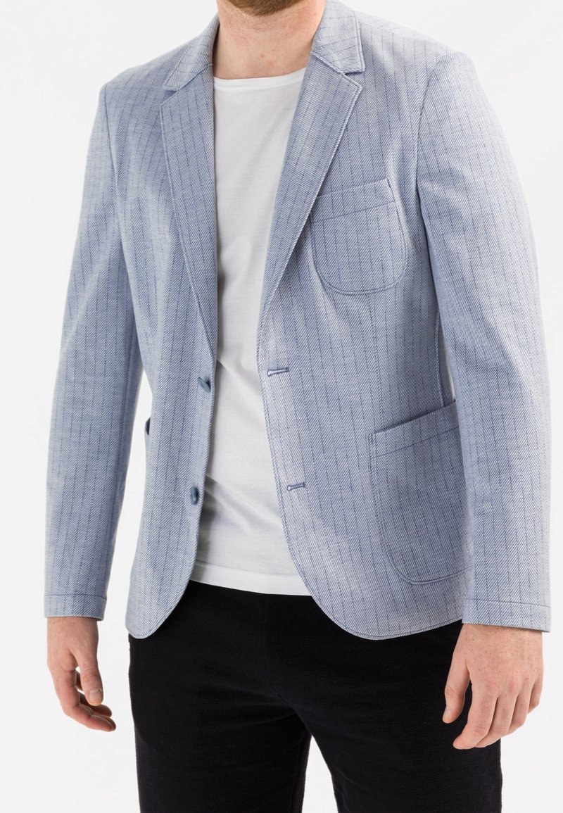 Homme portant un blazer bleu clair à fines rayures sur un T-shirt blanc et un pantalon noir sur fond blanc.