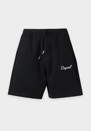 Νέος άντρας που φοράει κόκκινο t-shirt Dsquared2 Milano, μαύρες βερμούδες, λευκές κάλτσες και μαύρα αθλητικά παπούτσια Adidas, στέκεται μπροστά σε απλό φόντο.