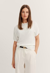 Weißes Poloshirt mit kurzen Ärmeln, weichem Material und geripptem Kragen, kombiniert mit hellen, gefältelten Hosen und einem schwarzen Gürtel.
