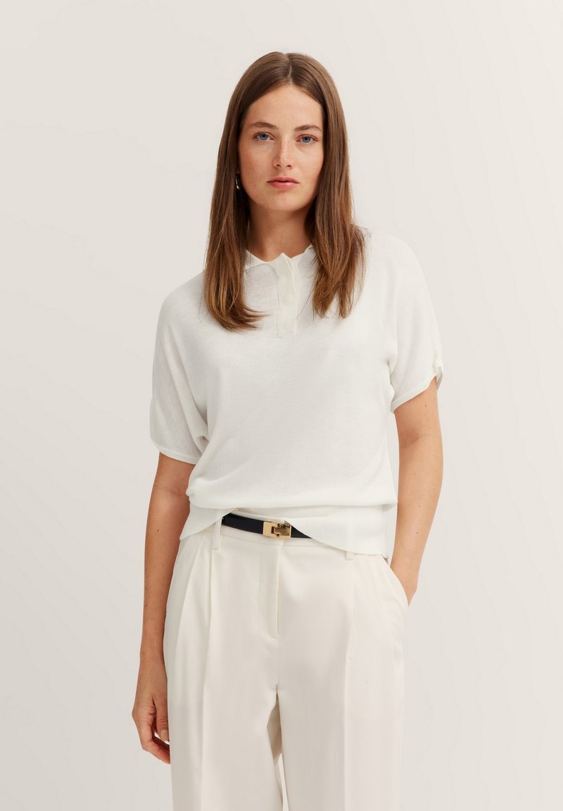 Weißes Poloshirt mit kurzen Ärmeln, weichem Material und geripptem Kragen, kombiniert mit hellen, gefältelten Hosen und einem schwarzen Gürtel.