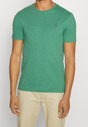 Homme portant un t-shirt vert à manches courtes avec un petit logo bleu brodé sur la poitrine, associé à un pantalon beige clair.