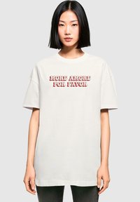 White oversized cotton t-shirt featuring red text that says "MORE AMORE POR FAVOR." Relaxed fit with dropped shoulders and short sleeves.