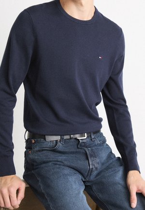 Mann trägt marineblauen Pullover mit langen Ärmeln und dunkelblaue Jeans, sitzt mit den Händen auf den Oberschenkeln vor weißem Hintergrund.